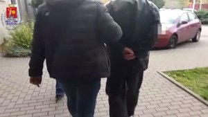 Zdjęcie przedstawia policjanta w ubraniu cywilnym, który prowadzi przez dziedziniec zatrzymanego mężczyznę ubranego na ciemno. Mężczyzna ten ma ręce z tyłu z założonymi kajdankami. Obok widać zaparkowany pojazd.