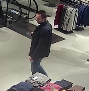 Zdjęcie przedstawia mężczyznę ubranego w ciemną kurtkę i ciemne spodnie. Stoi on w sklepie odzieżowym. Wokół widać stoliki  ułożoną odzieżą.