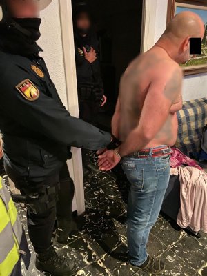 Zdjęcie przedstawia wnętrze pomieszczenia mieszkalnego, a w nim stojącego policjanta w granatowym mundurze, który przytrzymuje mężczyznę za ręce w kajdankach. Zatrzymany mężczyzna ma na sobie niebieskie spodnie jeansowe i ciemne klapki, pokryty jest licznymi tatuażami. W drzwiach do pomieszczenia stoi drugi policjant - również w granatowym mundurze.