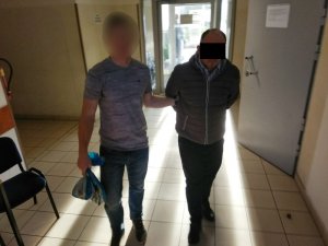 Zdjęcie przedstawia korytarz wewnątrz komendy. Widać na nim policjanta w ubraniu cywilnym (szara koszulka, niebieskie spodnie jeansowe, ciemne buty sportowe), który lewą ręką przytrzymuje zatrzymanego mężczyznę. W prawej ręce policjant trzyma dwie torebki foliowe w kolorze biało-niebieskim. Zatrzymany mężczyzna trzyma ręce za plecami. Ubrany jest w szarą kurtkę, spod której wystaje niebieska koszula, ciemne spodnie i ciemne buty. Twarz ma zasłoniętą czarnym paskiem. W tle widać szare drzwi prowadzące do pomieszczenia dla osób zatrzymanych i przeszklone drzwi wyjściowe.