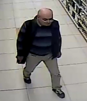 Zdjęcie przedstawia mężczyznę idącego wewnątrz sklepu, jest on w wieku około 50-60 lat, ma okulary na twarzy, ubrany jest w sweter w paski, ciemną kamizelkę, jasne spodnie. Mężczyzna posiada czarną torbę przewieszoną przez ramię. W tle widać półkę z asortymentem.