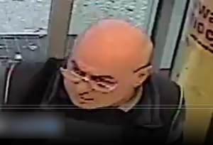 Zdjęcie przedstawia mężczyznę w wieku około 50-60 lat, ma okulary na twarzy, ubrany jest na ciemno. W tle widać drzwi przeszklone.