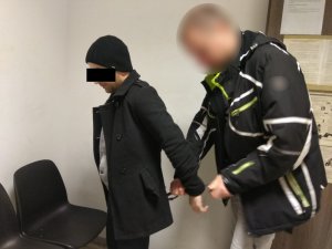 Zdjęcie przedstawia policjanta w ubraniu cywilnym, który zakłada kajdanki zatrzymanemu mężczyźnie na ręce trzymane z tyłu. Zatrzymany mężczyzna ubrany jest na ciemno, ma na głowie czarną czapkę. Obaj znajdują się w pomieszczeniu dla osób zatrzymanych.