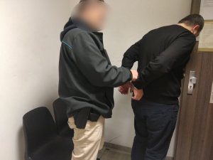 Zdjęcie przedstawia policjanta w umundurowaniu cywilnym, który zakłada kajdanki na ręce trzymane z tyłu zatrzymanemu mężczyźnie. Obaj znajdują się w pomieszczeniu dla osób zatrzymanych. Zatrzymany mężczyzna ubrany jest na ciemno.