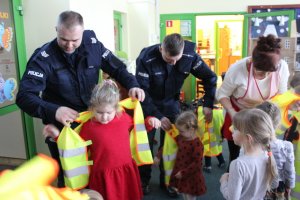 Zdjęcie przedstawia policjantów w granatowych mundurach, którzy pomagają zakładać dwóm dziewczynkom kamizelki odblaskowe. W tle widać inne dzieci.