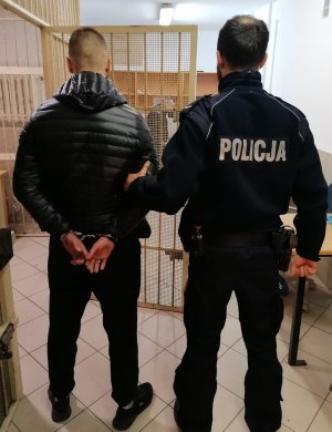 Na zdjęciu widać umundurowanego policjanta z zatrzymanym mężczyzną w kajdankach