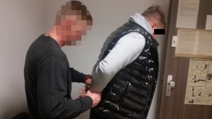 Zdjęcie przedstawia wnętrze pomieszczenia. Widać stojącego bokiem do obiektywu policjanta w cywilnym ubraniu, który nakłada kajdanki zatrzymanemu mężczyźnie. Mężczyzna ten ma szarą bluzę i czarny bezrękawnik. Policjant ubrany jest w ciemną bluzę.