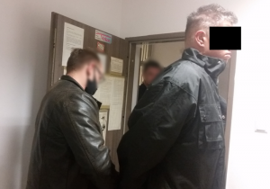 Zdjęcie przedstawia wnętrze pomieszczenia dla osób zatrzymanych. Widać na nim policjanta, w ubraniu cywilnym, który zakłada kajdanki stojącemu tyłem do niego mężczyźnie. Mężczyzna ten ma ciemną puchową kurtkę. Oczy zasłania mu czarny pasek