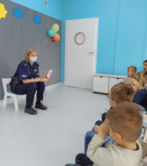 Zdjęcie przedstawia wnętrze sali przedszkolnej. Na krześle siedzi policjantka w granatowym mundurze, która ma maseczkę ochronną na twarzy. W ręku trzyma tarczę do zatrzymywania pojazdów. Przed nią w rzędzie siedzą dzieci.