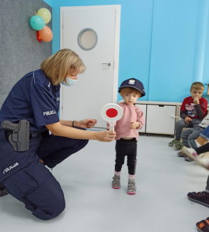 Zdjęcie przedstawia wnętrze sali przedszkolnej. Widać na nim policjantkę w granatowym mundurze, która ma maseczkę ochronną na twarzy. Pochyla się ona nad dzieckiem, które trzyma tarczę do zatrzymywania pojazdów. Dziewczynka ta ma założoną na głowę policyjną czapkę.