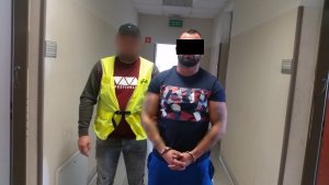 Zdjęcie przedstawia policjanta ubranego w odblaskową służbową kamizelkę, który przytrzymuje zatrzymanego mężczyznę. Górna część twarzy mężczyzny zasłonięta jest czarnym paskiem. Ma on na sobie koszulkę na krótki rękaw i niebieskie spodnie sportowe. Mężczyzna ten ma kajdanki założone na ręce trzymane z przodu. Obaj stoją na korytarzu, przodem do obiektywu.