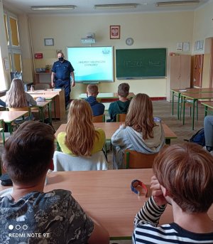 Zdjęcie przedstawia policjanta w umundurowaniu cywilnym, który stoi przed uczniami siedzącymi w ławkach. Za jego plecami, na tablicy jest wyświetlony slajd prezentacji.