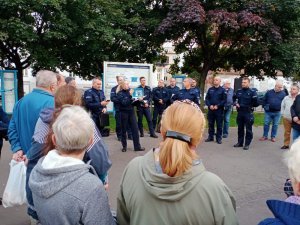 Zdjęcie przedstawia mieszkańców, którzy stoją na debacie. Przed nimi stoją policjanci w umundurowaniu służbowym. Spotkanie odbywa się na świeżym powietrzu.