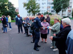 Zdjęcie przedstawia mieszkanki, którzy stoją na debacie. Przed nimi stoi policjant w umundurowaniu służbowym, który wręcza im ulotki. Spotkanie odbywa się na świeżym powietrzu.