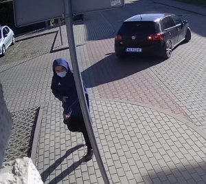 Zdjęcie przedstawia mężczyznę w wieku ok 30-35 lat, normalnej budowy ciała; ubrany w spodnie dresowe w kolorze czarnym z jasnymi szerokimi lampasami, obuwie sportowe w kolorze czarnym, bluzę z kapturem założonym na głowę w kolorze niebieskim, z białym paskiem na rękawach oraz białym znaczkiem na prawej piersi, na twarzy maseczka ochronna. W tle widać czarny samochód osobowy.