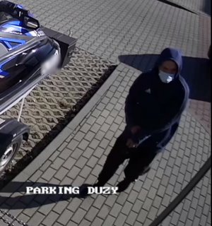 Zdjęcie przedstawia stojącego na chodniku mężczyznę. Jest on w wieku ok 30-35 lat, normalnej budowy ciała, ubrany w spodnie dresowe w kolorze czarnym z jasnymi szerokimi lampasami, obuwie sportowe w kolorze czarnym, bluzę z kapturem założonym na głowę w kolorze niebieskim, z białym paskiem na rękawach oraz białym znaczkiem na prawej piersi, na twarzy ma maseczkę ochronną.