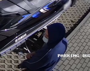 Zdjęcie przedstawia mężczyznę stojącego przy niebieskim skuterze. Ma on kaptur założony na głowę i maseczkę ochronną na twarzy.