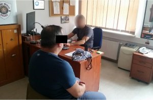 Zdjęcie przedstawia pokój służbowy, a w nim siedzącego za biurkiem policjanta w ubraniu cywilnym. Na wprost niego siedzi zatrzymany mężczyzna. Ma on granatową koszulkę. Siedzi tyłem do obiektywu.