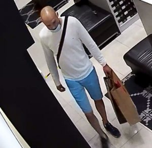 Zdjęcie przedstawia wnętrze sklepu, a w nim mężczyznę ubranego w białą bluzkę na długi rękaw, niebieskie spodenki do kolan, czarne buty, mężczyzna ma przewieszony przez klatkę piersiową czarny pasek, w lewej ręce trzyma papierową brązową torbę. Twarz zasłania mu jasna maseczka ochronna.
