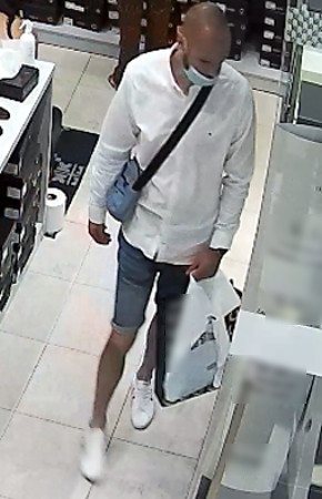 Zdjęcie przedstawia wnętrze sklepu, a w nim mężczyznę ubranego w białą koszulę na długi rękaw, niebieskie spodenki jeansowe do kolan, białe buty, mężczyzna ma przewieszony przez klatkę piersiową saszetkę błękitną na czarnym pasku, w lewej ręce trzyma torbę. Twarz zasłania mu jasna maseczka ochronna.
