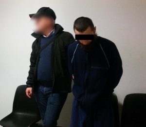 Zdjęcie przedstawia zatrzymanego mężczyznę, który ma twarz zasłoniętą twarz czarnym paskiem. Obok stoi policjant w ubraniu cywilnym, który przytrzymuje zatrzymanego mężczyznę.