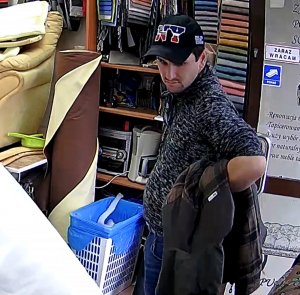 Zdjęcia przedstawia mężczyznę wewnątrz zakładu, ma on na sonie czapeczkę z daszkiem z napisem &quot;NY&quot;, szary sweter i granatowe spodnie jeansowe. Przez lewe przedramię ma przewieszoną ciemną kurtkę.
