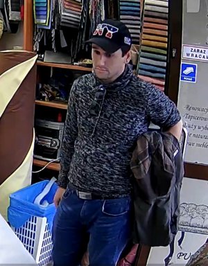 Zdjęcia przedstawia mężczyznę wewnątrz zakładu, ma on na sobie czapeczkę z daszkiem z napisem &quot;NY&quot;, szary sweter i granatowe spodnie jeansowe. Przez lewe przedramię ma przewieszoną ciemną kurtkę.