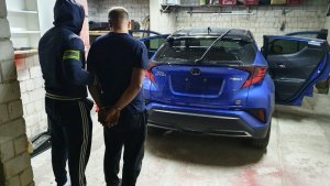 Zdjęcie przedstawia wnętrze pomieszczenia garażowego, a w nim stojącego w ubraniu cywilnym policjanta, który przytrzymuje za rękę zatrzymanego mężczyznę. W tle widać niebieski pojazd.