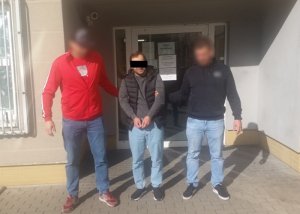 Zdjęcie przedstawia dwóch policjantów w ubraniach cywilnych, którzy stoją na zewnątrz, przed budynkiem. Obaj przytrzymują za ręce stojącego pomiędzy nimi zatrzymanego mężczyznę. Twarz zasłania mu czarny pasek.