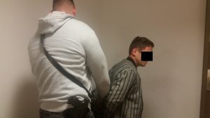 Zdjęcie przedstawia policjanta w ubraniu cywilnym, który zakłada kajdanki zatrzymanemu mężczyźnie. Policjant ma szarą bluzę.