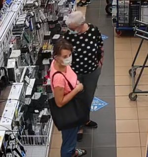Zdjęcie przedstawia dwie kobiety w sklepie. Jedna jest ubrana w czarną bluzkę w białe groszki, czarne spodnie i czarne buty, ma siwe, krótkie włosy; druga ma ciemne włosy, pomarańczową koszulkę na krótki rękaw, granatowe spodnie i klapki, trzyma na prawym ramieniu czarną torebkę. Obie mają maseczki ochronne na twarzy. Wokół nich widać półki z asortymentem sklepowym.