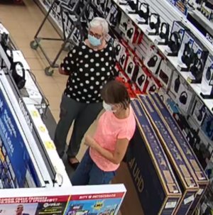 Zdjęcie przedstawia dwie kobiety w sklepie. Jedna jest ubrana w czarną bluzkę w białe groszki, czarne spodnie i czarne buty, ma siwe, krótkie włosy; druga ma ciemne włosy, pomarańczową koszulkę na krótki rękaw, granatowe spodnie. Obie mają maseczki ochronne na twarzy. Wokół nich widać półki z asortymentem sklepowym.