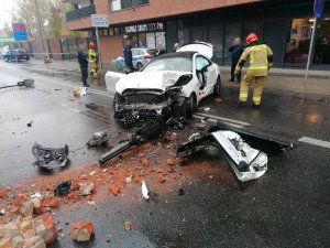 Zdjęcie przedstawia rozbity biały samochód. Wokół niego na ulicy stoją policjanci i strażacy.