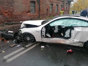 Zdjęcie przedstawia rozbity biały samochód. Wokół niego na ulicy stoją policjanci.