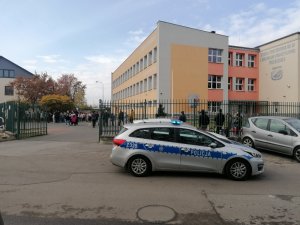 Zdjęcie przedstawia oznakowany policyjny radiowóz stojący na tle ogrodzenia szkolnego. Dalej widać uczestników ewakuacji.