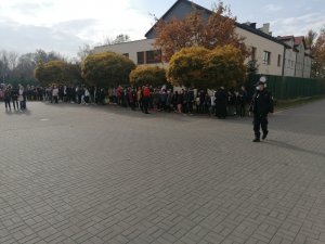 Zdjęcie przedstawia uczestników próbnej ewakuacji stojących na zewnątrz. Z prawej strony widać umundurowanego policjanta.