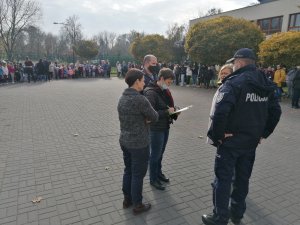 Zdjęcie przedstawia uczestników próbnej ewakuacji stojących na zewnątrz. Z prawej strony widać umundurowanego policjanta.