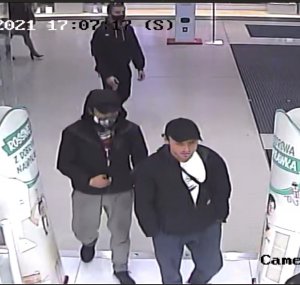 Zdjęcie przedstawia trzech mężczyzn wchodzących do sklepu. Pierwszy mężczyzna w wieku ok 30 lat, na głowie miał założoną czapkę z daszkiem w kolorze czarny, spodnie koloru czarnego, kurtkę w kolorze czarnym spod której wystawał biały t-shirt, przez tułów przewieszona tuba. Drugi z mężczyzn w wieku ok 25 lat ubrany w szare spodnie, kurtkę w kolorze czarnym, czarne obuwie sportowe, na głowie miał czarną czapkę z daszkiem a na nią zaciągnięty kaptur od kurtki, twarz osłonięta maseczką ochronną. Trzeci mężczyzna w wieku ok 25 lat, włosy ciemne krótkie, ubrany w czarną kurtkę z szarym kapturem, spodnie oraz buty w kolorze czarnym, na twarzy maseczka ochronna, na włosach okulary przeciwsłoneczne, miał ze sobą plecak.
