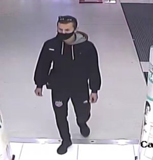 Zdjęcie przedstawia mężczyznę wewnątrz sklepu. Jest on w wieku ok 25vlat, włosy ciemne krótkie, ubrany w czarną kurtkę z szarym kapturem, spodnie oraz buty w kolorze czarnym, na twarzy maseczka ochronna, na włosach okulary przeciwsłoneczne, ma ze sobą plecak.
