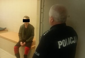 Zdjęcie przedstawia stojącego tyłem do obiektywu policjanta w t-shircie na krótki rękaw, który stoi w otwartych drzwiach policyjnej celi. W środku widać siedzącego mężczyznę. Jego prawie całą twarz zasłania czarny pasek.