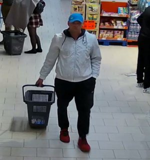 Zdjęcie przedstawia mężczyznę wewnątrz sklepu.  Jest to mężczyzna ok 35 lat, normalnej budowy ciała, wzrost ok 180cm, ubrany w kurtkę koloru białego, spodnie dresowe koloru czarnego, obuwie sportowe koloru czerwonego, czapka z daszkiem w kolorze niebieskim. W prawej ręce trzyma uchwyt wózka sklepowego. Lewą rękę trzyma w kieszeni spodni.
