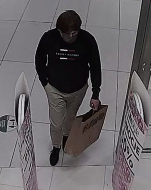 Zdjęcie przedstawia wchodzącego do sklepu mężczyznę w dłuższych włosach, ubranego w ciemną bluzę z napisem Tommy Hilfiger, jasne spodnie, buty koloru czarnego, maseczkę ochronną, w lewym ręku trzyma torbę papierową z logo sklepu „Smyk”. Prawą rękę mężczyzna trzyma w kieszeni spodni.