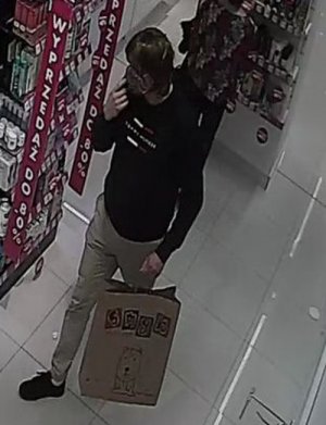 Zdjęcie przedstawia wchodzącego do sklepu mężczyznę w dłuższych włosach, ubranego w ciemną bluzę z napisem Tommy Hilfiger, jasne spodnie, buty koloru czarnego, maseczkę ochronną, w lewym ręku trzyma torbę papierową z logo sklepu „Smyk”. Prawą rękę mężczyzna trzyma w przy twarzy.