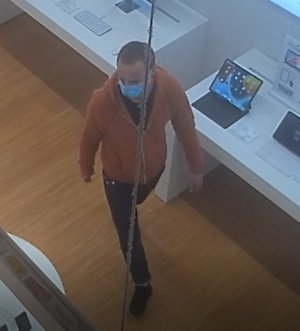 Zdjęcie przedstawia wnętrze sklepu, a w nim mężczyznę w wieku ok 30 lat,  wzrostu około 180 cm, ma on krótkie ciemne włosy, ubrany jest w rozpinaną bluzę w kolorze pomarańczowym, spodnie w kolorze czarnym , buty czarne, na twarzy ma maseczkę ochronną. W tle widać półkę, a którym stoi laptop.