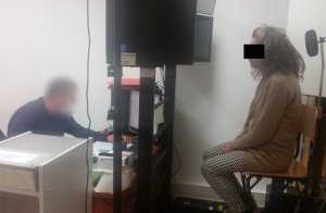 Zdjęcie przedstawia pokój służbowy. Po prawej stronie zdjęcia widać zatrzymaną kobietę, która siedzi na krześle lewym profilem do obiektywu aparatu. Ubrana jest w brązowy sweter i biało-czarne spodnie. Na wprost kobiety siedzi policjant w ubraniach cywilnych, za biurkiem, i obsługuje komputer.