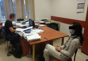 Zdjęcie przedstawia kobietę w szarym dresie, która siedzi przy biurku. Jest ona skierowana lewym profilem do aparatu. Ma ciemne włosy, na twarzy ma błękitną maseczkę ochronną. Na rękach trzymanych z przodu ma założone kajdanki. Przy biurku, na którym leżą teczki z dokumentami siedzi policjant przed komputerem.