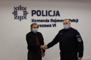 Zdjęcie przedstawia policjanta w umundurowaniu służbowym, który podaje rękę mężczyźnie. Obaj stoją na tle białej ściany, gdzie widnieje napis Komenda Rejonowa Policji Warszawa VI.
