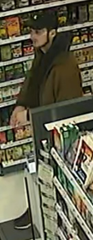 Zdjęcie przedstawia mężczyznę wewnątrz sklepu. Jest to mężczyzna w wieku około 25 lat, około 180 cm wzrostu, w czarnej czapce z daszkiem z białym elementem nad daszkiem, ubrany w ciemną kurtkę, czarną bluzę, na twarzy widoczny charakterystyczny ciemny zarost w formie szpiczastej bródki.