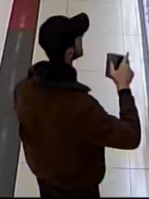Zdjęcie przedstawia mężczyznę wewnątrz sklepu. Jest to mężczyzna w czarnej czapce z daszkiem, ubrany w ciemną kurtkę, czarne spodnie, na twarzy widoczny charakterystyczny ciemny zarost. W prawej ręce trzyma jakiś czarny przedmiot.