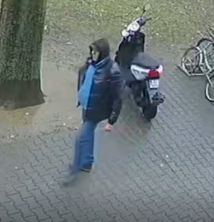 Zdjęcie przedstawia mężczyznę, który ma na sobie ciemną kurtkę, niebieską bluzkę, niebieskie spodnie jeansowe. Na głowie ma założony kaptur. W tle widać zaparkowany skuter.